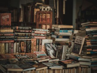 Ein unordentlich aufgestapelter Haufen Bücher vor einem Regal in dem weitere Bücher stehen.