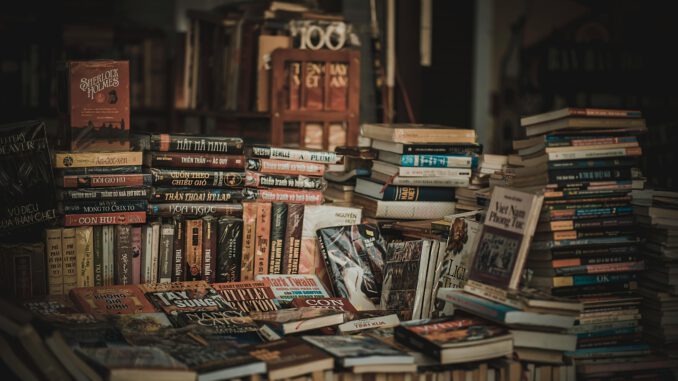 Ein unordentlich aufgestapelter Haufen Bücher vor einem Regal in dem weitere Bücher stehen.